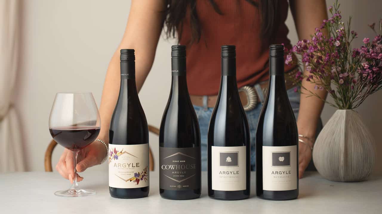 Four bottles of Argyle wine in a line placed on a table next to a glass of red wine. A server stands behind them waiting to pour.