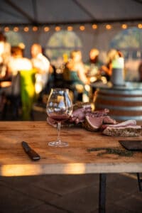 A glass of pinot noir on a butcher block with cuts of meat and guests dining in the Chef's Garden tent in the background.