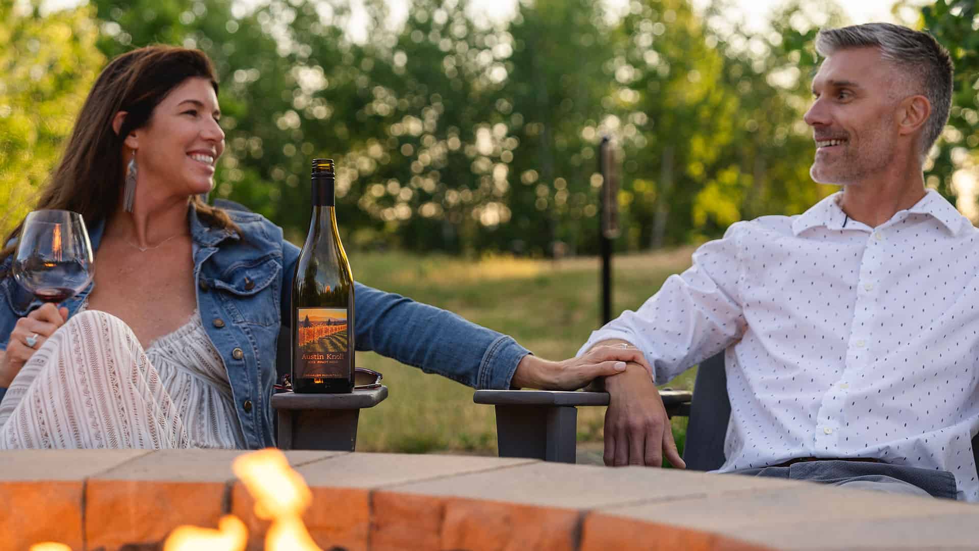A couple sitting by the fire pit drinking Austin Knoll Pinot Noir.