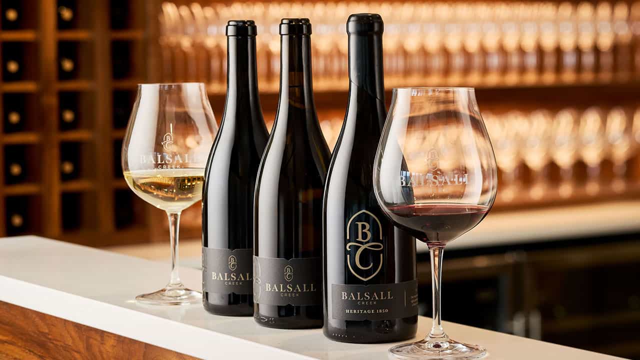 Three bottles of Balsall Creek wine next two to wine glasses on a countertop. A lit shelf of wine glasses in the background.