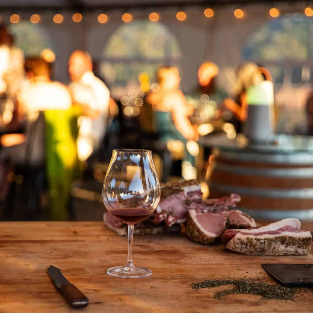 A glass of pinot noir on a butcher block with cuts of meat and guests dining in the Chef's Garden tent in the background.
