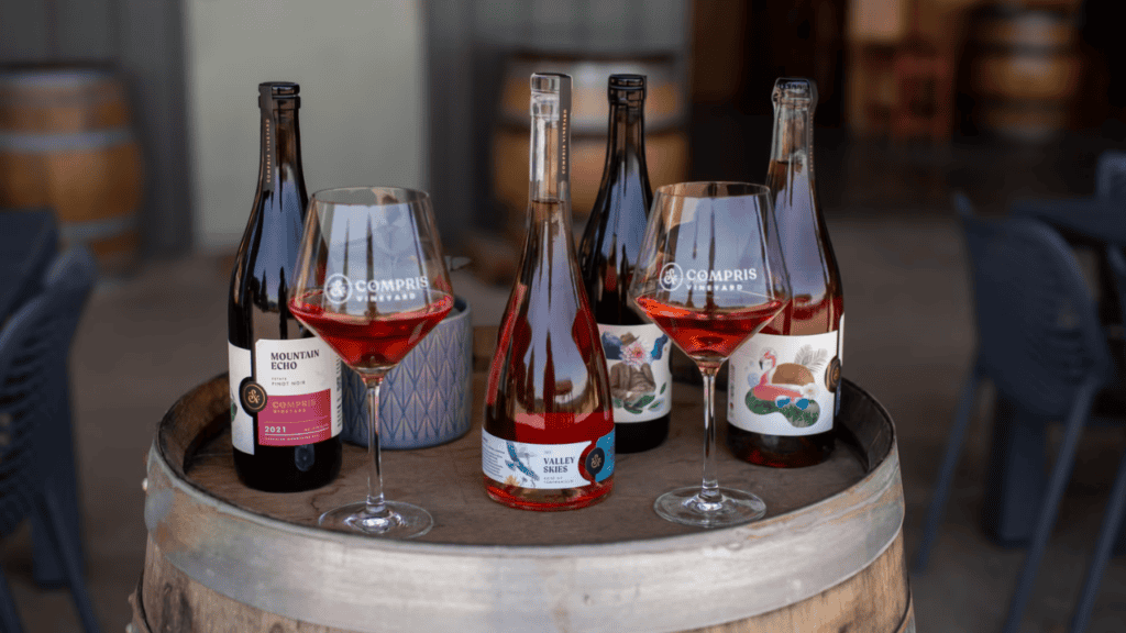Four bottles of wine from Compris Vineyard and two wine glasses on a wine barrel.