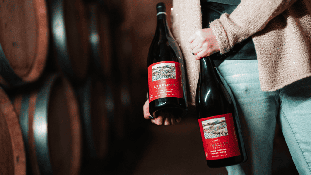 A woman holding two bottles of Lemelson Vineyard wine near wine barrels.