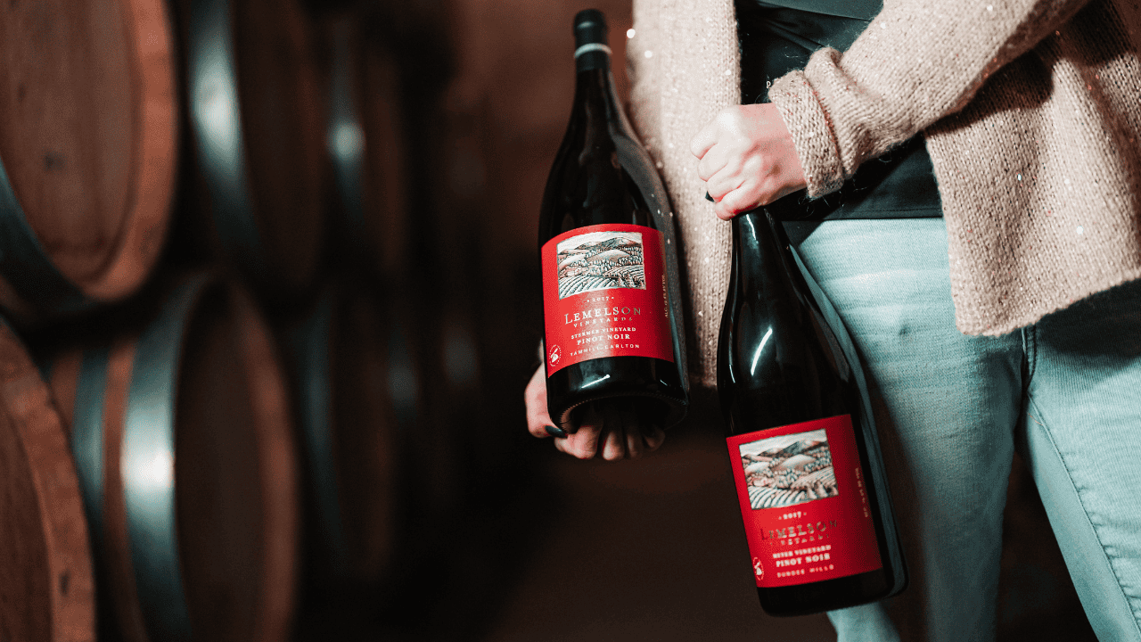 A woman holding two bottles of Lemelson Vineyard wine near wine barrels.