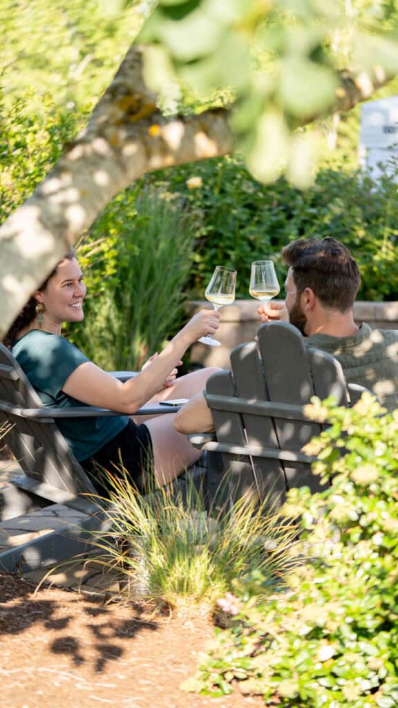 A couple sitting in chairs around the firepit, drinking white wine.